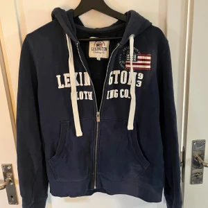 Mörkblå hoodie från Lexington - Säljer en snygg mörkblå hoodie från Lexington med dragkedja och vit text på framsidan. Den har en najs luva med tjocka vita snören och en cool amerikansk flagga som detalj. Perfekt för en avslappnad stil!                                      Vid andra frågor, tveka inte att höra av dig🙏🏼😊