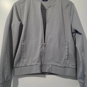 Grå jacka från GANT oanvänd  - Snygg grå jacka från GANT med dragkedja och ribbade muddar. Jackan har ett subtilt mönster och två praktiska fickor framtill. Perfekt för en stilren look.