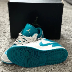 Nike Air Jordan 1 low aquatone - Snygga Nike Air Jordans i en fräsch kombination av vitt och turkos. Skorna har klassisk snörning och en stilren design med det ikoniska Jordan-emblemet på plösen. Perfekta för dig som vill ha en sportig och trendig look.