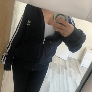 Svart träningsjacka från Adidas - Säljer en svart träningsjacka från Adidas med de klassiska tre vita ränderna längs ärmarna. Jackan har dragkedja framtill och en broderad logga på bröstet. Perfekt för en sportig look!