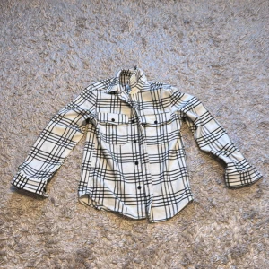 Vit och svart rutig skjorta - Snygg vit och svart rutig skjorta med långa ärmar och knappar framtill. Skjortan har två bröstfickor med knappar och en klassisk krage. Perfekt för en stilren look.