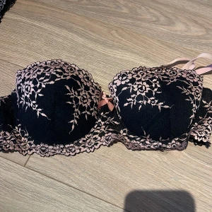 Svart och rosa spets-bh - Elegant svart bh med rosa spetsdetaljer och en söt rosett framtill. Den har vadderade kupor och justerbara axelband för bästa passform. Perfekt för att känna sig extra fin.
