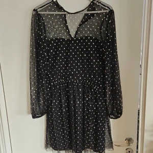 Svart klänning med prickar - Snygg svart klänning med genomskinliga långa ärmar och ett prickigt mönster i guld. Klänningen har en v-ringad design och ett elegant fall. Perfekt för en kväll ute eller en speciell tillställning.