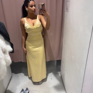 Gul långklänning med knyt - Snygg gul långklänning med knyt i ryggen och v-ringad design. Perfekt för sommarens festligheter. Klänningen har en elegant passform och är ärmlös, vilket ger en stilren look.