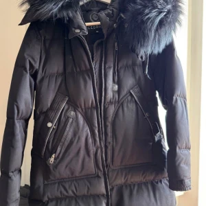 Svart dunjacka med pälsdetalj från Cedrico - Snygg svart dunjacka från Cedrico med en äkta stor pälsdetalj på huvan. Jackan har dragkedja och knappar framtill samt praktiska fickor med dragkedja. Perfekt för kalla dagar.