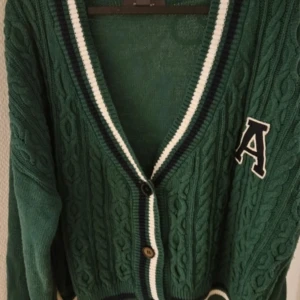 Grön kofta från Primark - Säljer en snygg grön kofta från Primark med kabelstickat mönster och en stor bokstav 'A' på bröstet. Koftan har en v-ringad design med knappar framtill och ribbade kanter i vitt och mörkblått.