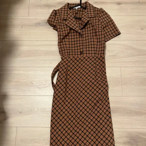 Rutig klänning med bälte - Snygg rutig klänning i brunt och svart med korta ärmar och knappar framtill. Den har en klassisk krage och ett matchande bälte i midjan för en fin silhuett. Perfekt för en stilren look!