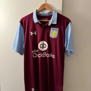 Aston Villa - Snygg vinröd fotbollströja med ljusblå ärmar och krage från Under Armour. Tröjan har AVFC-logga på bröstet och QuickBooks som sponsor. Perfekt för fotbollsfans som vill visa sitt stöd med stil.