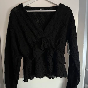 Svart blus från Gina Tricot - Snygg svart blus från Gina Tricot med volangdetaljer och v-ringning. Blusen har långa ärmar och en texturerad yta som ger en elegant touch. Passar till både vardags och fest. Aldrig använd men finns ingen lapp då den redan var borta i butiken. Storlek M. Priser går att diskuteras 🩷