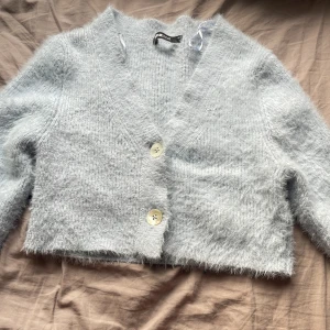 Ljusblå fluffig kofta från Gina Tricot - Mysig ljusblå kofta från Gina Tricot i fluffigt material. Den har en croppad passform med knappar framtill och långa ärmar. Perfekt för kyliga dagar när du vill ha något bekvämt och stilrent. Använd 1 gång, jättefin till layering