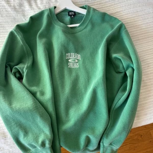 Grön sweatshirt från Urban outfitters - Säljer en grön sweatshirt från Urban outfitters med texten 'Colorado Springs 1995' tryckt på bröstet. Tröjan har en klassisk rund hals och långa ärmar. Perfekt för en avslappnad stil.