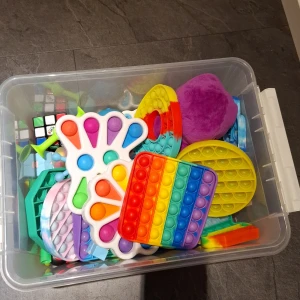 Samling av pop it-leksaker i olika former och färger - Stor samling av pop it-leksaker i en genomskinlig plastlåda. Innehåller olika former som fyrkantiga, runda och hjärtformade i regnbågsfärger, gult och blått. Perfekt för stressavlastning och sensorisk lek. Inkluderar även en Rubiks kub och andra små fidget-leksaker.