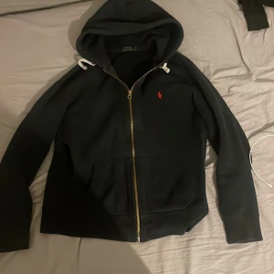 Svart hoodie från Polo Ralph Lauren - Säljer en stilren svart hoodie från Polo Ralph Lauren med dragkedja och huva. Den har en liten röd logotyp på bröstet och vita snören vid huvan. Perfekt för en avslappnad look.