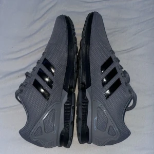 Grå sneakers från Adidas - Snygga grå sneakers från Adidas med svarta detaljer och den klassiska tre-randiga designen på sidan. Skorna har en modern look med en robust sula och är perfekta för en sportig stil. Skorna i nypris kostar 1200kr och skorna är i bra skick men lite sliten sula bara och nån skråma på själva skorna‼️