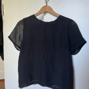 Svart blus från Jeane Blush - Snygg svart blus från Jeane Blush med korta ärmar och transparenta detaljer vid axlarna. Perfekt för en stilren look. Blusen har en enkel design med en knapp i nacken för enkel på- och avtagning. Storlek M, men passar även S och Xs