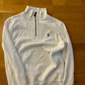 Vit tröja med dragkedja från Ralph Lauren - Säljer en stilren vit tröja från Ralph Lauren med halv dragkedja och broderad logga på bröstet. Tröjan har långa ärmar och en hög krage, perfekt för kyligare dagar.