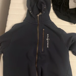 Mörkblå jacka med huva från Sail Racing - Säljer en mörkblå jacka med huva från Sail Racing. Jackan har en dragkedja framtill och en bröstficka med dragkedja. Perfekt för kyliga dagar med sin stilrena design och funktionella detaljer.