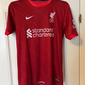 Röd Liverpool fotbollströja från Nike - Säljer en röd Liverpool fotbollströja från Nike med vita detaljer och klubbens emblem på bröstet. Tröjan har korta ärmar och ett stilrent mönster. Perfekt för alla Liverpool-fans! 🔴⚽