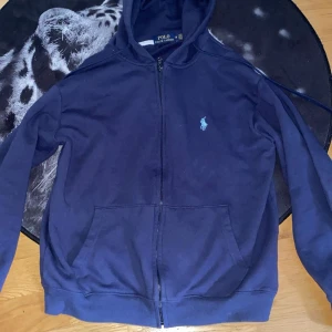 Blå hoodie från Ralph Lauren - Säljer en snygg blå hoodie från Ralph Lauren med dragkedja och det klassiska broderade logotypen på bröstet. Tröjan har en bekväm passform och är perfekt för en avslappnad stil. Den har också praktiska fickor framtill. Nypris 2295kr säljer för 450kr. Använd tiotal gånger 