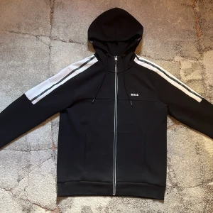 Hugo boss  - Snygg svart hoodie från BOSS med dragkedja och vita ränder längs ärmarna. Perfekt för en stilren outfit, köptes från Johnells för 1900 förra månaden och har knappt använts. Tveka inte på frågor, svarar snabbt