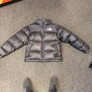 Svart dunjacka från The North Face - Säljer en svart dunjacka från The North Face med dragkedja och hög krage. Jackan har en pufferdesign och är perfekt för kalla dagar. Den har långa ärmar och en justerbar huva för extra skydd mot väder och vind. Storlek S