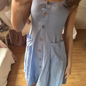 Blå randig klänning med knappar - Säljer en söt blå randig klänning med knappar framtill och fickor på sidorna. Klänningen har en kropps passform och är perfekt för varma dagar. 