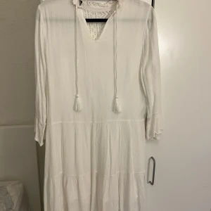 Vit klänning med spetsdetaljer - Vit klänning med spetsdetaljer på överdelen och knytband vid halsen. Klänningen har trekvartsärmar och en luftig passform. Oanvänd!
