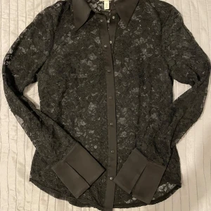 Svart spetsblus från H&M - Snygg svart spetsblus från H&M med långa ärmar och knappar framtill. Blusen har eleganta manschetter och en klassisk krage. Perfekt för en stilren look.