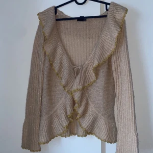 Beige stickad kofta med volangdetaljer - Säljer en superfin beige stickad kofta med glittriga inslag och volangdetaljer. Koftan har långa ärmar och knytning framtill. Helt perfekt till en tråkig outfit och passformen på koftan är väldigt smickrande!😊