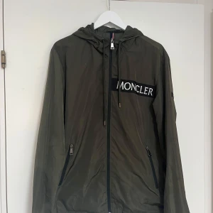 Grön vindjacka från Moncler - Säljer en snygg grön vindjacka från Moncler med dragkedja och huva. Jackan har Moncler-logga på bröstet och en cool serietidningsdetalj på insidan. Perfekt för höst och vår! Storlek L men passar M också.