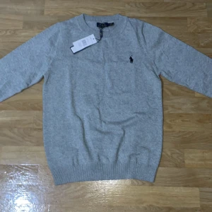 Grå tröja från Polo Ralph Lauren - Säljer en stilren grå tröja från Polo Ralph Lauren med det klassiska broderade logotypen på bröstet. Tröjan har långa ärmar och ribbade muddar vid ärmslut och nederkant. Perfekt för en avslappnad och snygg look.