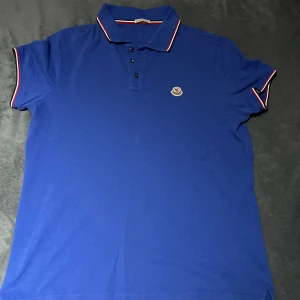 Blå pikétröja från Moncler - Snygg blå pikétröja från Moncler med klassisk krage och knappar. Tröjan har korta ärmar med röd och vit randdetalj vid ärmslut och krage. Perfekt för en stilren look.