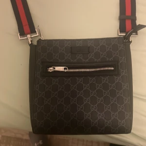 Svart axelväska från Gucci riktigt snygg väska - Snygg svart axelväska från Gucci med det klassiska GG-mönstret. Väskan har en justerbar axelrem i rött och svart samt en praktisk dragkedja på framsidan. Perfekt för att bära dina nödvändigheter med stil.