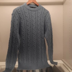 Blå stickad tröja från 157 - Säljer en snygg blå stickad tröja från 157. Tröjan har ett klassiskt flätmönster och rund halsringning. Perfekt för kyligare dagar med sin mysiga och stilrena design. Passar bra till både jeans och kjol.