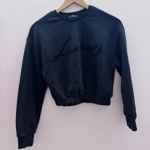 Svart croppad sweatshirt  - Säljer en svart croppad sweatshirt med texten 'Live happy' på framsidan. Tröjan har långa ärmar och en elastisk nederkant för en bekväm passform. Perfekt för en avslappnad stil.