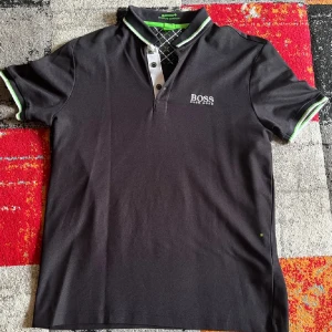 Svart kortärmad pikétröja från Boss - Säljer en snygg svart pikétröja från Hugo Boss med gröna och vita detaljer på kragen och ärmsluten. Storlek M och normal i storlek! Hör av er vid frågor eller funderingar.