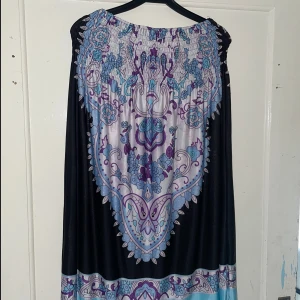 Mönstrad klänning med blommor - Säljer en superfin klänning med ett färgglatt blommönster i lila, blått och svart. Klänningen har en lös passform och är perfekt för sommarens alla fester. Den har en smockad överdel som ger en bekväm passform. Perfekt för en bohemisk look!