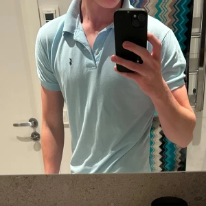 Ljusblå pikétröja från Ralph Lauren - Säljer en snygg ljusblå pikétröja från Ralph Lauren. Den har en klassisk krage. Perfekt för både vardag och lite finare tillfällen. Tröjan är i bra skick och har den ikoniska loggan på bröstet. Säljer då den e lite för liten. Modell 182 väger ca 70