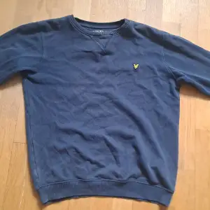 Säljer en snygg blå tröja från Lyle & Scott. Den har en klassisk design med ett gult emblem på bröstet. Tröjan är i bomull och har långa ärmar. Perfekt för höst och vår! Observera att det finns ett litet hål på ena ärmen som går att fixa.