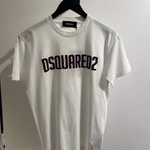 Vit t-shirt från Dsquared2 - Säljer en helt ny vit t-shirt från Dsquared2 med deras ikoniska logga tryckt i svart och rött på bröstet. T-shirten är kortärmad och gjord i mjuk bomull, perfekt för en casual look. Passar både till vardags och fest!