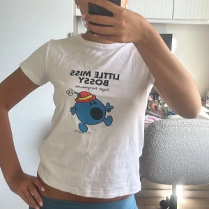 Vit t-shirt med Little Miss Bossy-tryck - Säljer en superfin vit t-shirt med ett tryck av Little Miss Bossy i blått och rött. T-shirten är kortärmad och har en normal passform. Perfekt för en avslappnad look eller som en rolig detalj i din outfit!