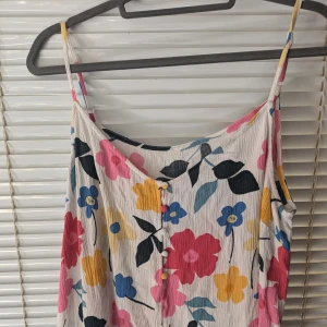 Blommig sommarklänning med knappar - Säljer en superfin blommig sommarklänning med smala axelband. Klänningen har ett färgglatt mönster med stora blommor i rosa, gul, blå och röd på en vit bakgrund. Den har knappar framtill som en söt detalj.Storlek XL, men storlekslapp klippte bort