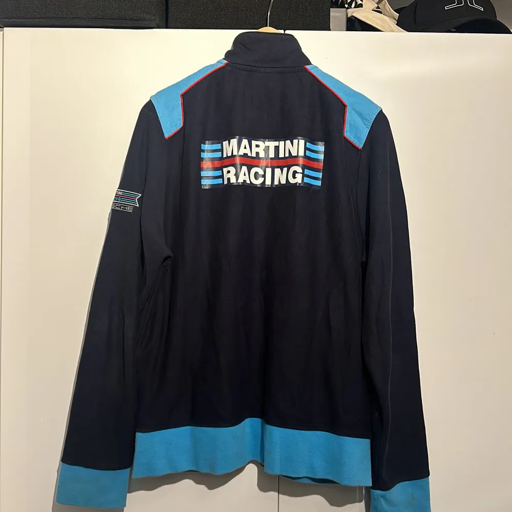 Säljer en snygg blå Cardigan från Porsche Design med coola Martini Racing detaljer. Tröjan har en dragkedja framtill och är långärmad. Den har ett broderat nummer '1' på framsidan och texten 'MARTINI RACING' på baksidan. Perfekt för höst och vår!. Tröjor & Koftor.