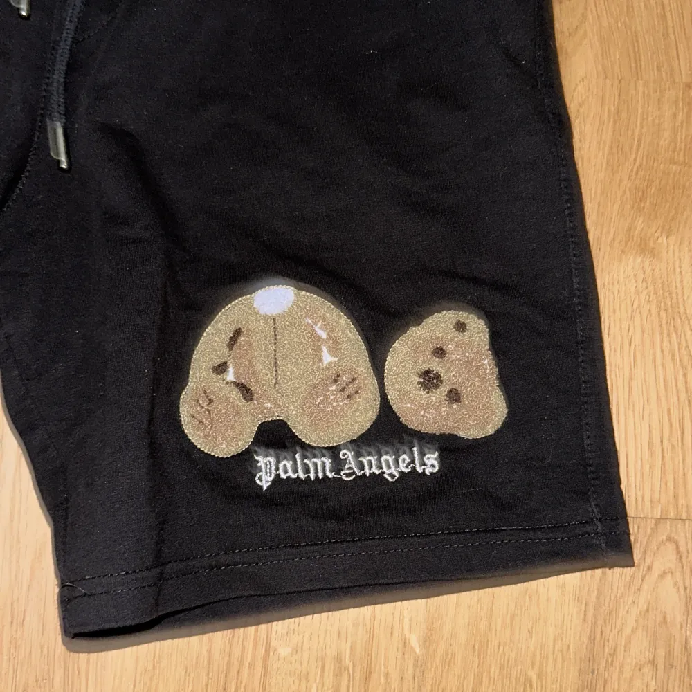 Säljer ett par svarta shorts från Palm Angels. De är i bra skick och har en cool design med en nallebrodyr på framsidan. Shortsen har en elastisk midja med dragsko för bästa passform. Perfekta för en avslappnad stil!. Shorts.