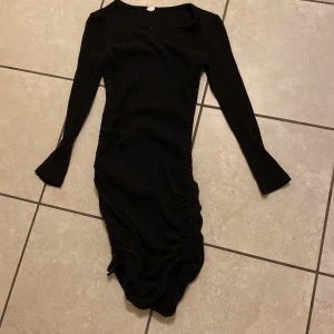 Svart ribbad klänning - Säljer en snygg svart ribbad klänning med lång ärm och en cool v-ringad detalj. Klänningen har en tight passform och är perfekt för festliga tillfällen. Den har också en snygg rynkad detalj längs sidorna som ger en extra touch. Perfekt för höst och vinter!