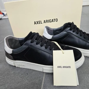 Svarta sneakers från Axel Arigato - Säljer ett par stilrena svarta sneakers från Axel Arigato. Skorna har en snygg kombination av svart och vit färg med en vit sula. De är tillverkade i skinn och har en klassisk snörning. Perfekta för både vardag och fest!