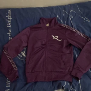 Lila jacka från Rocawear - Säljer en snygg lila tröja från Rocawear i storlek Xs/XXs. Jackan har en dragkedja framtill och långa ärmar. Den är dekorerad med ett stilrent broderat 'R' på bröstet och längs ärmarna. Perfekt för en cool och avslappnad stil!