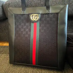 Säljer en snygg svart axelväska från Gucci med det klassiska GG-mönstret. Väskan har en elegant design med gröna och röda ränder i mitten och ett stort GG-emblem i guld. Den är tillverkad i skinn och har en praktisk dragkedja upptill. Mycket rymlig!