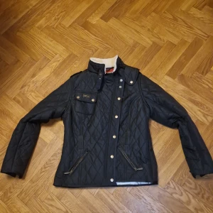 Svart quiltad jacka från Barbour - Snygg svart quiltad jacka från Barbour med dragkedja och knappar. Jackan har en unik innerfoderdesign med karta och är perfekt för höst och vår. Den har långa ärmar och en hög krage med mjukt foder.
