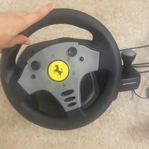 Ratt och pedaler från Thrustmaster med Ferrari-logga - Säljer en ratt och pedalset från Thrustmaster med en ikonisk Ferrari-logga i mitten. Ratten har en svart och grå design med flera knappar för spelkontroll. Pedalerna är svarta och robusta, perfekta för racingentusiaster. Perfekt för en realistisk körupplevelse i dina favoritspel! Endast för PC